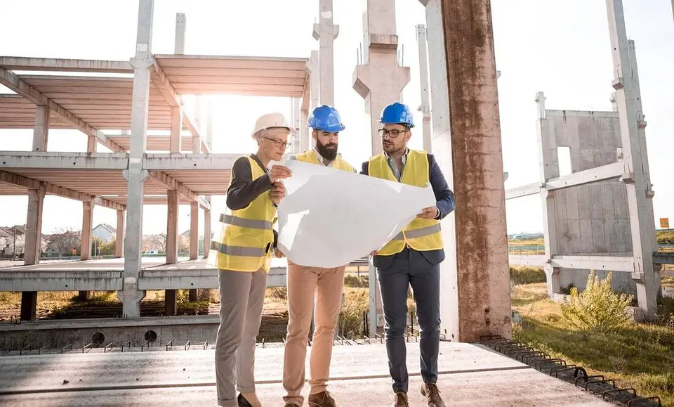 Three construction professionals in hard hats and safety vests reviewing plans together on a job site